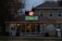 A florist shop with a prominently displayed neon sign reading 'Art Floral' in pink and green atop the entrance. The storefront windows display various plants and floral arrangements. The building has a quaint, cozy appearance, set against a backdrop of trees and a slightly overcast sky. Warm orange string lights are strung along the roofline, adding a welcoming touch.