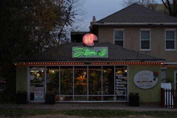 A florist shop with a prominently displayed neon sign reading 'Art Floral' in pink and green atop the entrance. The storefront windows display various plants and floral arrangements. The building has a quaint, cozy appearance, set against a backdrop of trees and a slightly overcast sky. Warm orange string lights are strung along the roofline, adding a welcoming touch.