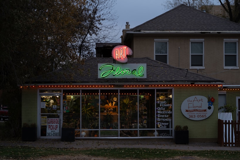 A florist shop with a prominently displayed neon sign reading 'Art Floral' in pink and green atop the entrance. The storefront windows display various plants and floral arrangements. The building has a quaint, cozy appearance, set against a backdrop of trees and a slightly overcast sky. Warm orange string lights are strung along the roofline, adding a welcoming touch.