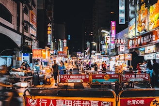 A bustling night market scene with numerous food stalls under vibrant, illuminated signs. The street is crowded with people exploring the various vendors. Brightly lit signage in multiple colors decorates the market, creating a lively atmosphere.