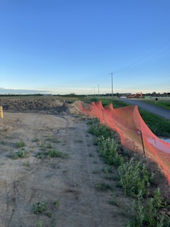 A construction site bordered by an orange net fence with visible piles of dirt. The area is positioned alongside a road and features green grass and vegetation. Utility poles with power lines run parallel to the road, and the sky is clear with a gradient of blue.