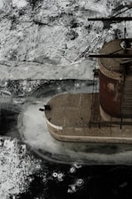 An industrial structure, resembling a silo or storage container, is partially visible, surrounded by a frozen surface. The ice shows cracks and patterns with varying shades of white and gray. The structure appears rusty, indicating weathering and time.