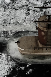An industrial structure, resembling a silo or storage container, is partially visible, surrounded by a frozen surface. The ice shows cracks and patterns with varying shades of white and gray. The structure appears rusty, indicating weathering and time.