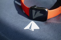 A smartwatch with an orange band is placed on top of a dark fabric surface that has a white arrow symbol printed on it.