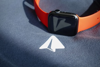 A smartwatch with an orange band is placed on top of a dark fabric surface that has a white arrow symbol printed on it.