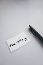 A black pen lies on a smooth, light-colored surface next to a white index card with the handwritten message 'stay healthy'.