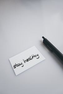A black pen lies on a smooth, light-colored surface next to a white index card with the handwritten message 'stay healthy'.