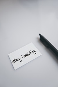 A black pen lies on a smooth, light-colored surface next to a white index card with the handwritten message 'stay healthy'.