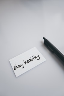A black pen lies on a smooth, light-colored surface next to a white index card with the handwritten message 'stay healthy'.