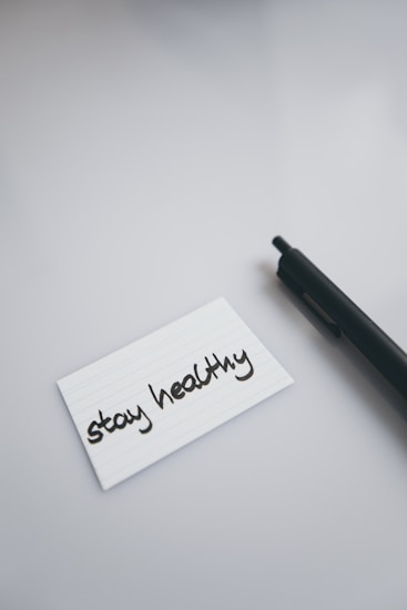A black pen lies on a smooth, light-colored surface next to a white index card with the handwritten message 'stay healthy'.