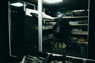 A person wearing a hooded jacket is browsing shelves of produce in a dimly lit grocery store. The shelves are stocked with various fruits and vegetables, and the lighting creates a moody, subdued atmosphere.