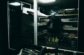 A person wearing a hooded jacket is browsing shelves of produce in a dimly lit grocery store. The shelves are stocked with various fruits and vegetables, and the lighting creates a moody, subdued atmosphere.