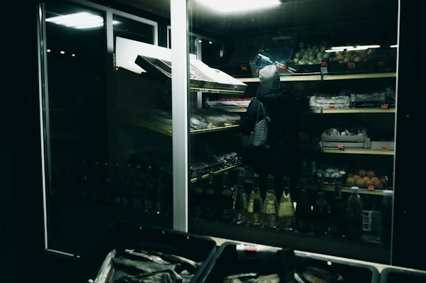 A person wearing a hooded jacket is browsing shelves of produce in a dimly lit grocery store. The shelves are stocked with various fruits and vegetables, and the lighting creates a moody, subdued atmosphere.