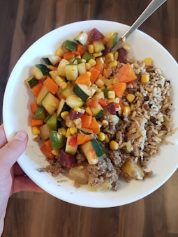 A white plate holds a colorful mix of diced vegetables including zucchini, corn, carrots, and onions, accompanied by minced meat and a serving of rice with grains.
