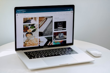 A laptop displaying a blog or webpage with images and text, sitting on a white table accompanied by a wireless mouse.