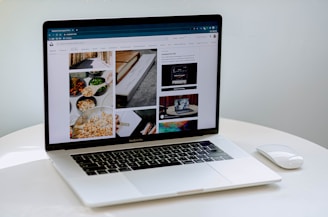 A laptop displaying a blog or webpage with images and text, sitting on a white table accompanied by a wireless mouse.
