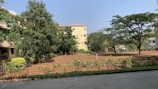 A serene outdoor scene featuring a large open yard with bare soil surrounded by greenery. Several trees provide a lush canopy, and there are well-maintained shrubs and hedges along a metal fence. In the background, residential buildings with light-colored walls and red tile roofs are partially obscured by the trees.