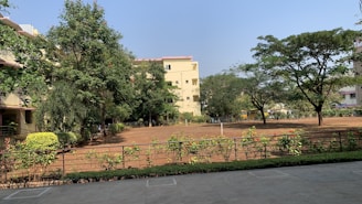 A serene outdoor scene featuring a large open yard with bare soil surrounded by greenery. Several trees provide a lush canopy, and there are well-maintained shrubs and hedges along a metal fence. In the background, residential buildings with light-colored walls and red tile roofs are partially obscured by the trees.