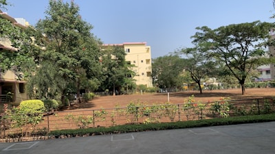 A serene outdoor scene featuring a large open yard with bare soil surrounded by greenery. Several trees provide a lush canopy, and there are well-maintained shrubs and hedges along a metal fence. In the background, residential buildings with light-colored walls and red tile roofs are partially obscured by the trees.