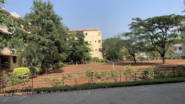 A serene outdoor scene featuring a large open yard with bare soil surrounded by greenery. Several trees provide a lush canopy, and there are well-maintained shrubs and hedges along a metal fence. In the background, residential buildings with light-colored walls and red tile roofs are partially obscured by the trees.