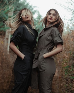 Two individuals stand outdoors, each wearing a stylish jumpsuit. One wears a black jumpsuit, and the other wears an olive green one. Their hair is slightly windswept, adding a dynamic feel to the image. The background features blurred greenery, possibly a forested area with an overcast sky.