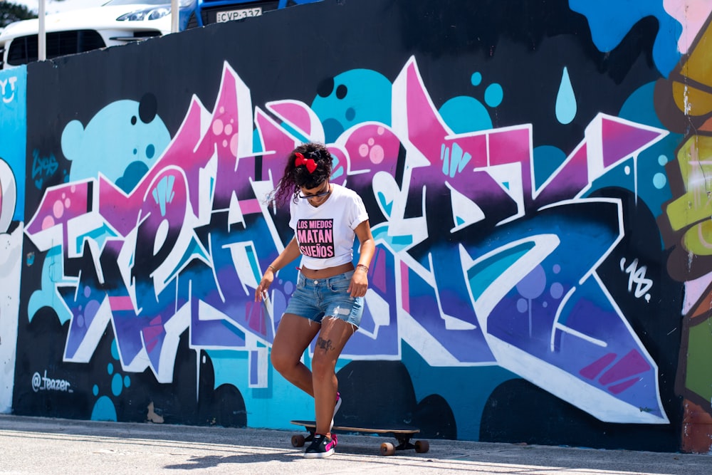 woman in white t-shirt and blue denim shorts standing beside wall with graffiti during daytime