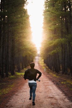 A person is running down a forest path surrounded by tall trees. The path is earthy and stretches into the distance, where sunlight streams through the trees.
