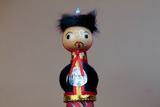 A wooden doll features a round head with painted facial expressions, including eyes and a small mustache. It wears a traditional outfit with a black coat, red and gold accents, and a small silver medallion. The doll's head is adorned with a tuft of black, furry material resembling hair.