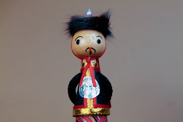 A wooden doll features a round head with painted facial expressions, including eyes and a small mustache. It wears a traditional outfit with a black coat, red and gold accents, and a small silver medallion. The doll's head is adorned with a tuft of black, furry material resembling hair.