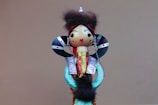 A wooden doll with a painted face, wearing a colorful outfit and adorned with a fur trim. The doll has large, decorative black and colorful earrings, with a red and black striped headpiece. The body is dressed in ornate, colorful materials, including blue, gold, and red patterns, giving it an intricate and traditional appearance.