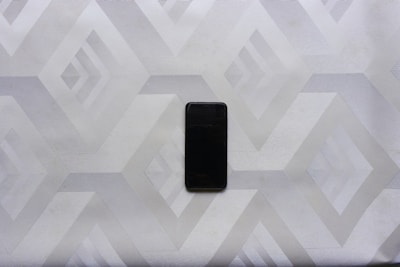 A smartphone lies on a surface with a geometric, diamond-patterned design. The phone is centrally placed, and the overall tone is a mixture of light grays and whites, creating a minimalistic and modern aesthetic.