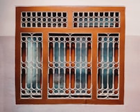 A window with a decorative metal grille featuring circular and arched patterns. The frame is painted brown, and the wall surrounding it is painted in two-tone colors, comprising a light beige and a darker shade.