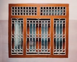 A window with a decorative metal grille featuring circular and arched patterns. The frame is painted brown, and the wall surrounding it is painted in two-tone colors, comprising a light beige and a darker shade.