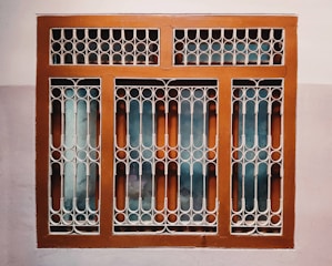 A window with a decorative metal grille featuring circular and arched patterns. The frame is painted brown, and the wall surrounding it is painted in two-tone colors, comprising a light beige and a darker shade.