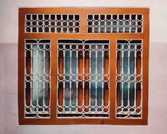 A window with a decorative metal grille featuring circular and arched patterns. The frame is painted brown, and the wall surrounding it is painted in two-tone colors, comprising a light beige and a darker shade.