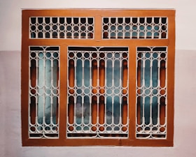 A window with a decorative metal grille featuring circular and arched patterns. The frame is painted brown, and the wall surrounding it is painted in two-tone colors, comprising a light beige and a darker shade.