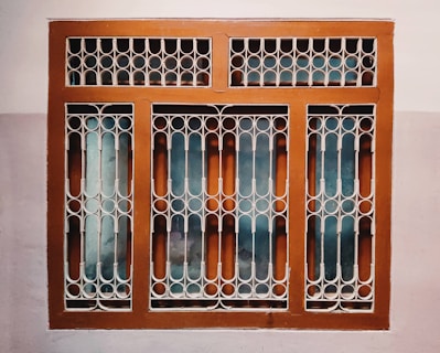 A window with a decorative metal grille featuring circular and arched patterns. The frame is painted brown, and the wall surrounding it is painted in two-tone colors, comprising a light beige and a darker shade.