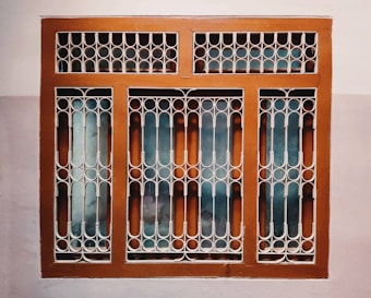 A window with a decorative metal grille featuring circular and arched patterns. The frame is painted brown, and the wall surrounding it is painted in two-tone colors, comprising a light beige and a darker shade.