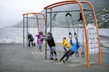 Several people are exercising at an outdoor gym near a body of water. The gym features orange and black equipment suitable for different workout routines. The background shows a scenic view of mountains and greenery across the water. The individuals, wearing athletic clothing, appear to be engaged in various exercises.