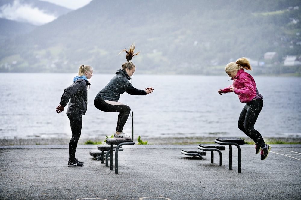 woman in pink jacket and black pants jumping on black bench near body of water during