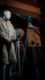 A man wearing a fur coat stands beside a cinematographic setup, including a vintage camera on a tripod and studio lighting equipment. The setting is dimly lit, creating a dramatic atmosphere.