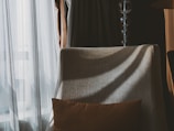 A cozy interior scene featuring a beige upholstered chair positioned next to a sheer curtain with sunlight streaming through. A decorative brown pillow rests on the chair, and a modern standing lamp with spherical glass elements is visible behind it. Shadows are cast across the surface, adding depth and texture.
