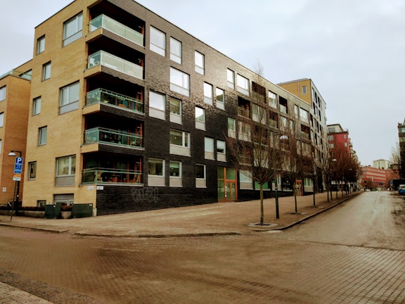 A modern apartment building with a combination of beige and dark brick exterior, featuring large windows and balconies with glass railings. The building is situated on a quiet, empty street lined with bare trees, indicating a winter or early spring season.