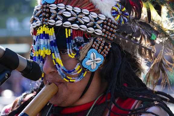 A person wearing a colorful and ornate headdress made of beads, shells, and feathers plays a wooden wind instrument. The headdress includes intricate patterns with blue, yellow, white, and brown elements, alongside feathers in various colors.