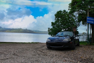A black convertible sports car is parked on a gravel surface beside a serene lake. The sky features a mix of clouds and blue patches, while lush green hills are visible across the water. A large tree stands to the right of the car, and a sign pointing to a trattoria is also present.