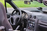 The interior of a convertible car features a black leather steering wheel with a Mazda logo. The dashboard includes round air vents and an audio system with various dials and controls. In the background, a grassy field can be seen through the windshield.
