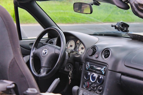 The interior of a convertible car features a black leather steering wheel with a Mazda logo. The dashboard includes round air vents and an audio system with various dials and controls. In the background, a grassy field can be seen through the windshield.