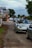 A line of cars, mostly convertibles, is parked along a road. The lead car is a silver convertible with the driver visible, wearing a cap. There are signposts on the left, advertising local businesses, and trees stand in the background against a cloudy sky.