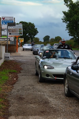 A line of cars, mostly convertibles, is parked along a road. The lead car is a silver convertible with the driver visible, wearing a cap. There are signposts on the left, advertising local businesses, and trees stand in the background against a cloudy sky.