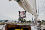 A pirate flag featuring a skull and crossbones design with a red bandana is prominently displayed on a sailboat. The background includes a marina with several buildings, trees, and distant hills under a cloudy sky. Part of the boat's fixtures, such as ropes and a winch, are visible in the foreground.